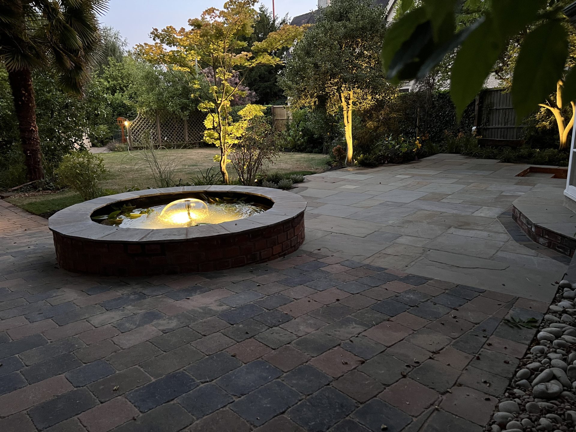 Garden water feature and tree uplighting on a block-paved patio at dusk